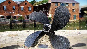 En stor, svart skulptur i form av en propeller står på en offentlig plats, och en plakett finns placerad i närheten. I bakgrunden syns en byggnad i tegel med texten "QUAYARTS", och framför den ligger en kanal med vatten och svanar.