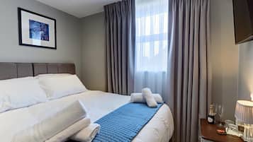 Quarto de hotel com cama, mesa de cabeceira com abajur e janela com cortinas.