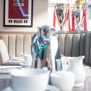 Un coin repas avec une table dressée pour deux personnes, une affiche encadrée du Liverpool F.C. et des trophées de la Premier League exposés sur une étagère.
