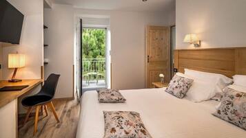 Une chambre d’hôtel comprenant un lit, un bureau avec une chaise, une télévision, une armoire en bois et un balcon donnant sur un paysage verdoyant.
