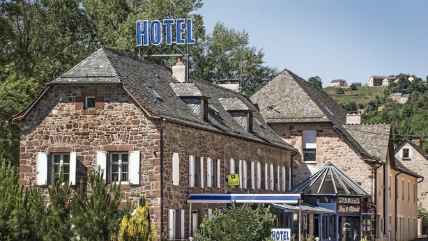 Un edificio de piedra con un letrero que dice 'HOTEL', un pequeño anexo con toldo azul y una colina al fondo.