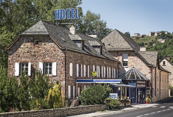 Un edificio de piedra con un letrero que dice 'HOTEL', un pequeño anexo con toldo azul y una colina al fondo.