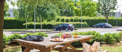 Una mesa de picnic con comida y bebidas, rodeada de follaje y automóviles estacionados.