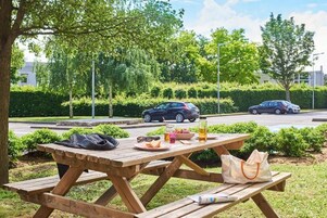 Una mesa de picnic con comida y bebidas, rodeada de follaje y automóviles estacionados.