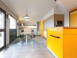 Un área de recepción de hotel moderna con un mostrador amarillo brillante, un cartel que dice 'Bienvenue' y un espacio de descanso con mesas y sillas redondas.