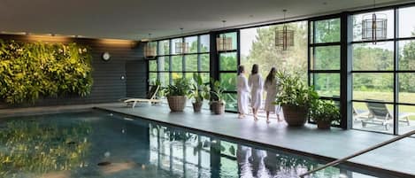 Une piscine intérieure dotée de grandes fenêtres, de plantes en pot et offrant une vue sur la verdure environnante.