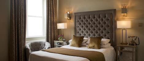 A hotel room with a large bed, a tufted headboard, two bedside lamps, a nightstand with a lamp, a chair, and a window with curtains.