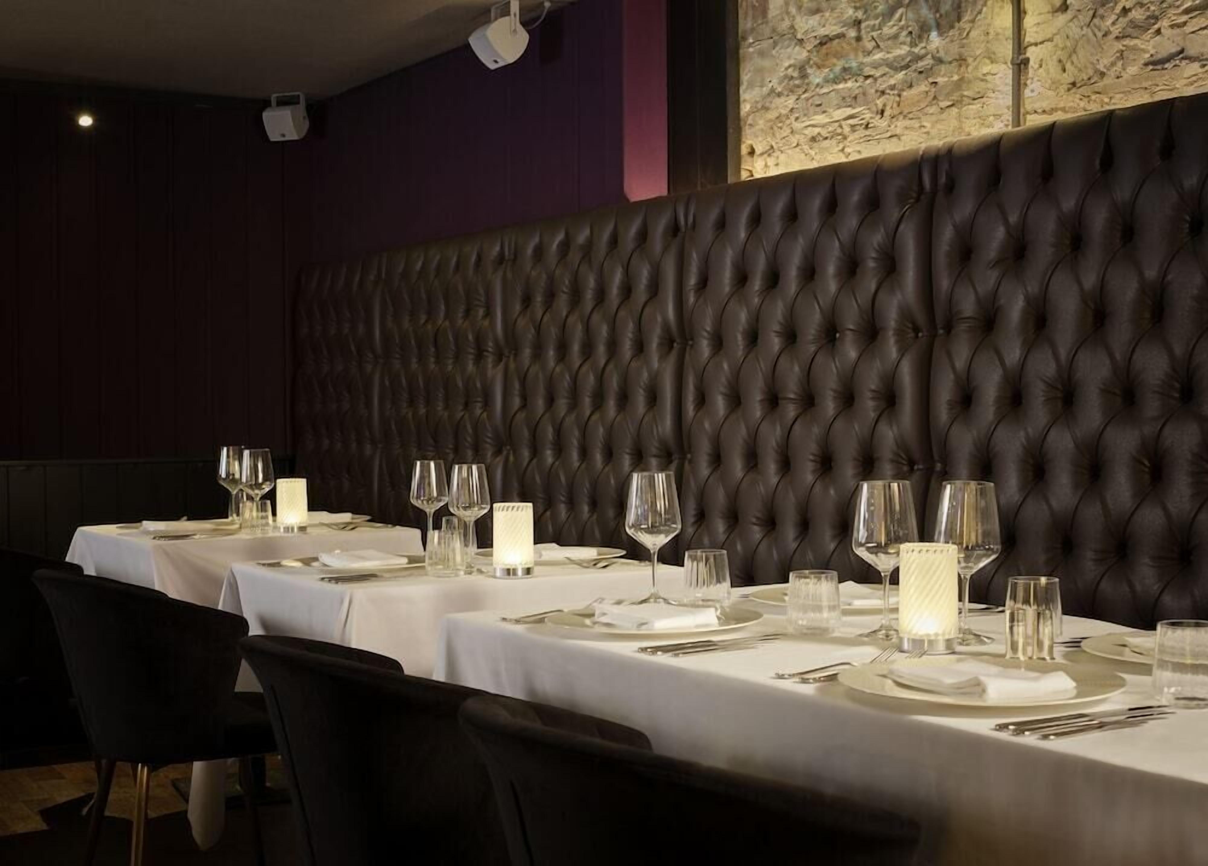 Un restaurante con una pared larga, oscura y con textura, manteles blancos y centros de mesa con velas.