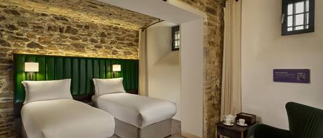 A room with two beds, a green upholstered headboard, a small table with a teapot, and a green armchair.