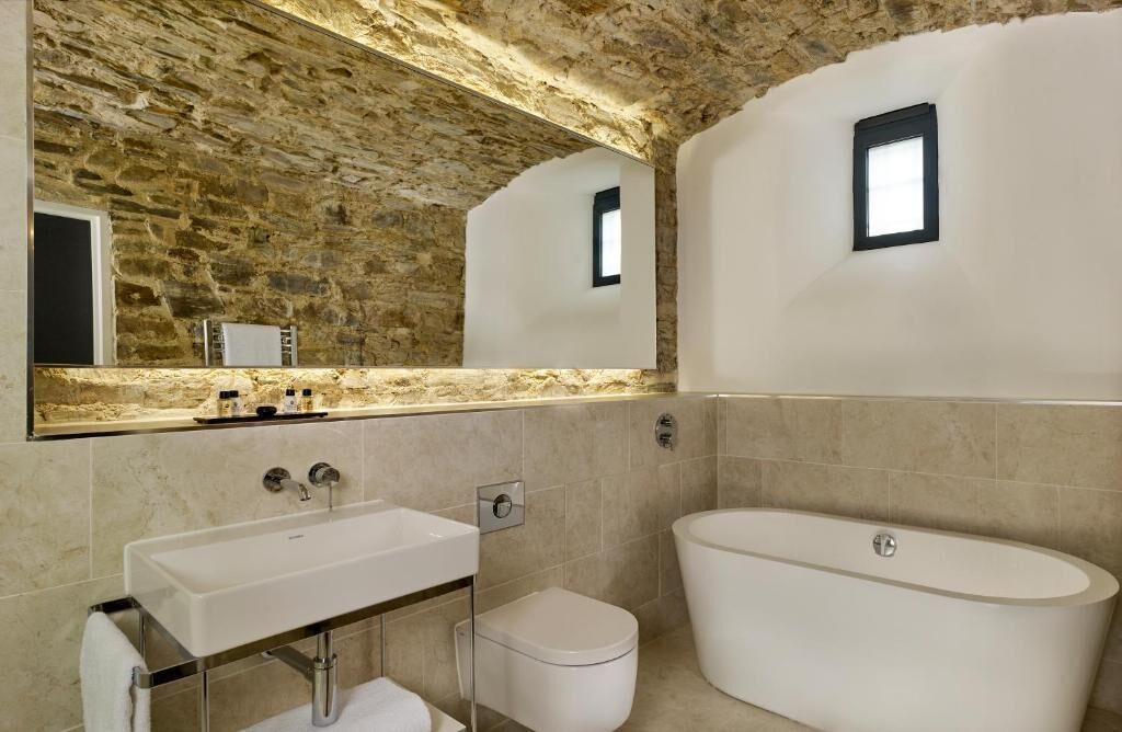 A bathroom with a stone wall, a large mirror, a sink, a toilet, and a bathtub.