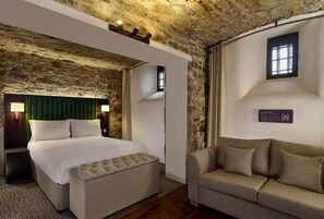 Un dormitorio con pared de piedra, una cama con cabecera verde, una mesita de noche, un sofá y una ventana pequeña.