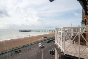 Vista a la playa desde un balcón, con un muelle a lo lejos, un estacionamiento y una barandilla blanca.