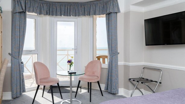 A hotel room with a balcony, two pink chairs, a small round table, a TV mounted on the wall, and a metal drying rack. - The Lanes Hotel (Brighton)