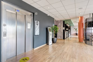 Un pasillo interior con mostrador de recepción, panel digital de ascensor y una planta en maceta.