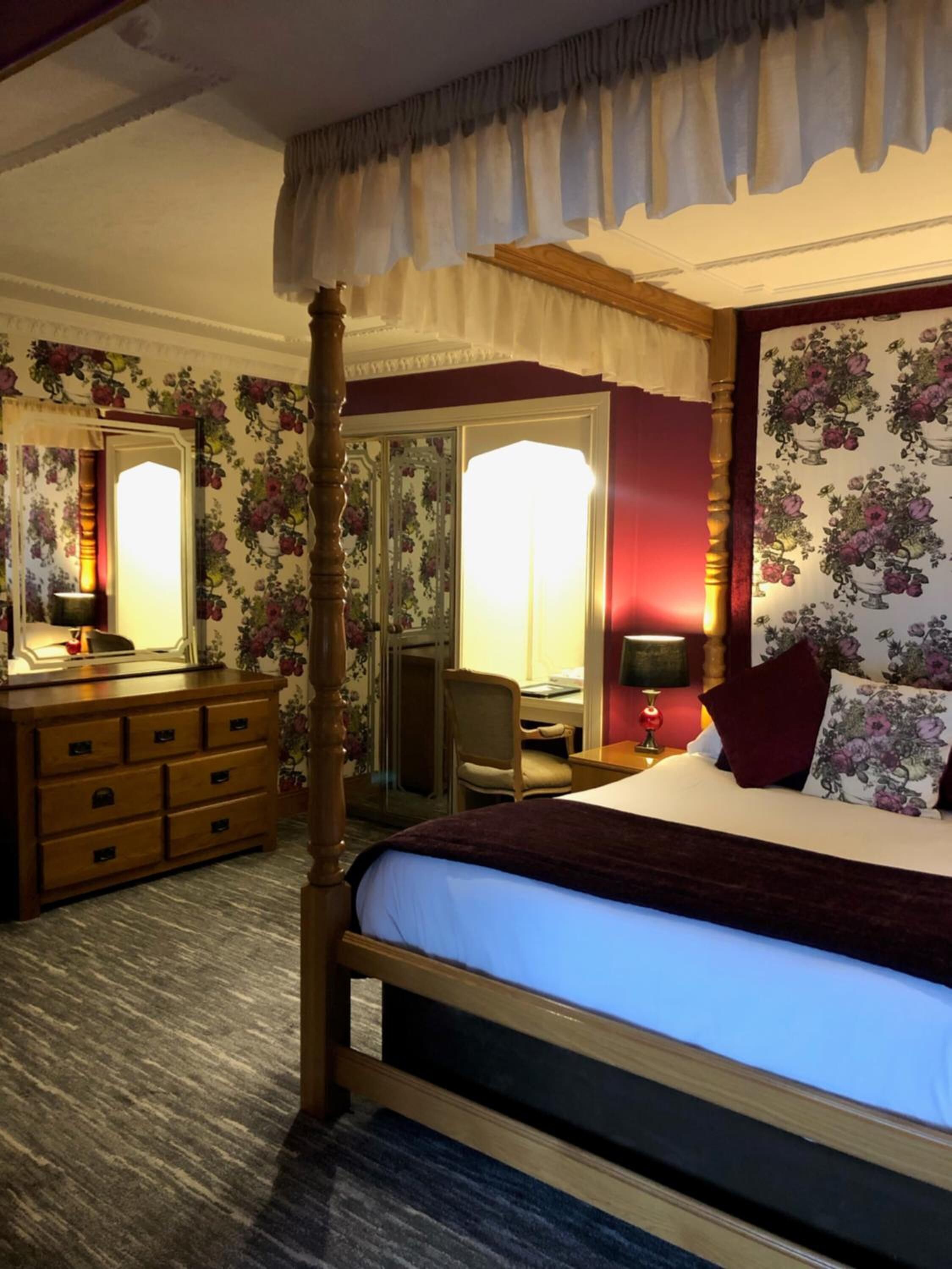 Un lit à baldaquin avec une tête de lit sombre, une commode en bois dotée de nombreux tiroirs, un miroir et une chaise.