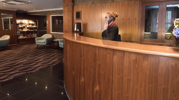 A reception area with a wooden counter, a seated guest area with chairs, a patterned carpet, and a wall-mounted television. - Palace Hotel (Peterhead)