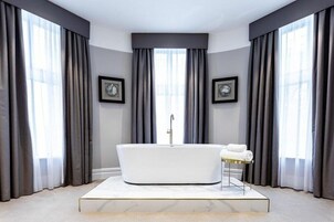 Ein modernes Badezimmer mit einer freistehenden Badewanne, großen Fenstern mit grauen Vorhängen und zwei gerahmten Uhren an den Wänden.