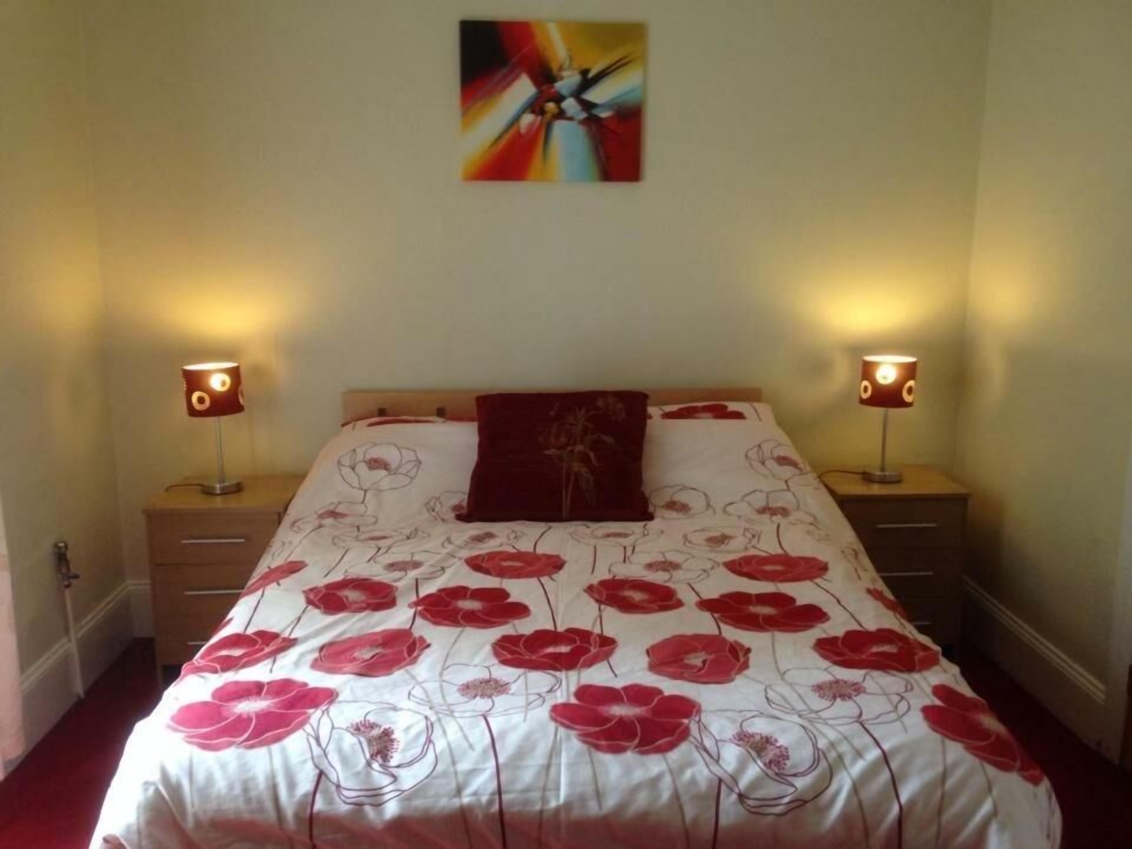 A bedroom with a bed, two bedside lamps, a wall art piece, and a floral-patterned bedspread.