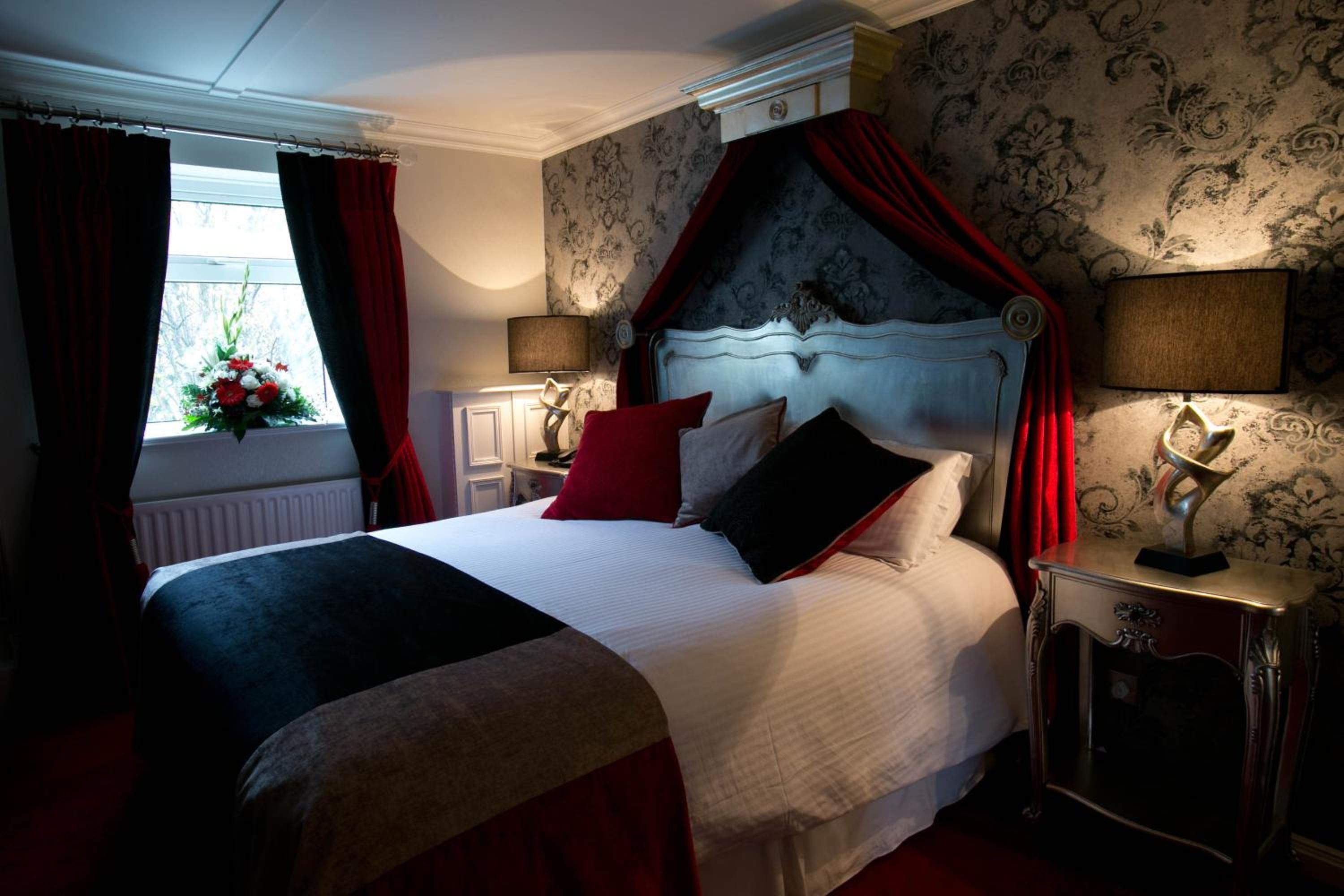 A bedroom with a four-poster bed, a window with curtains, a bedside table with a lamp, and a floral arrangement on the windowsill.