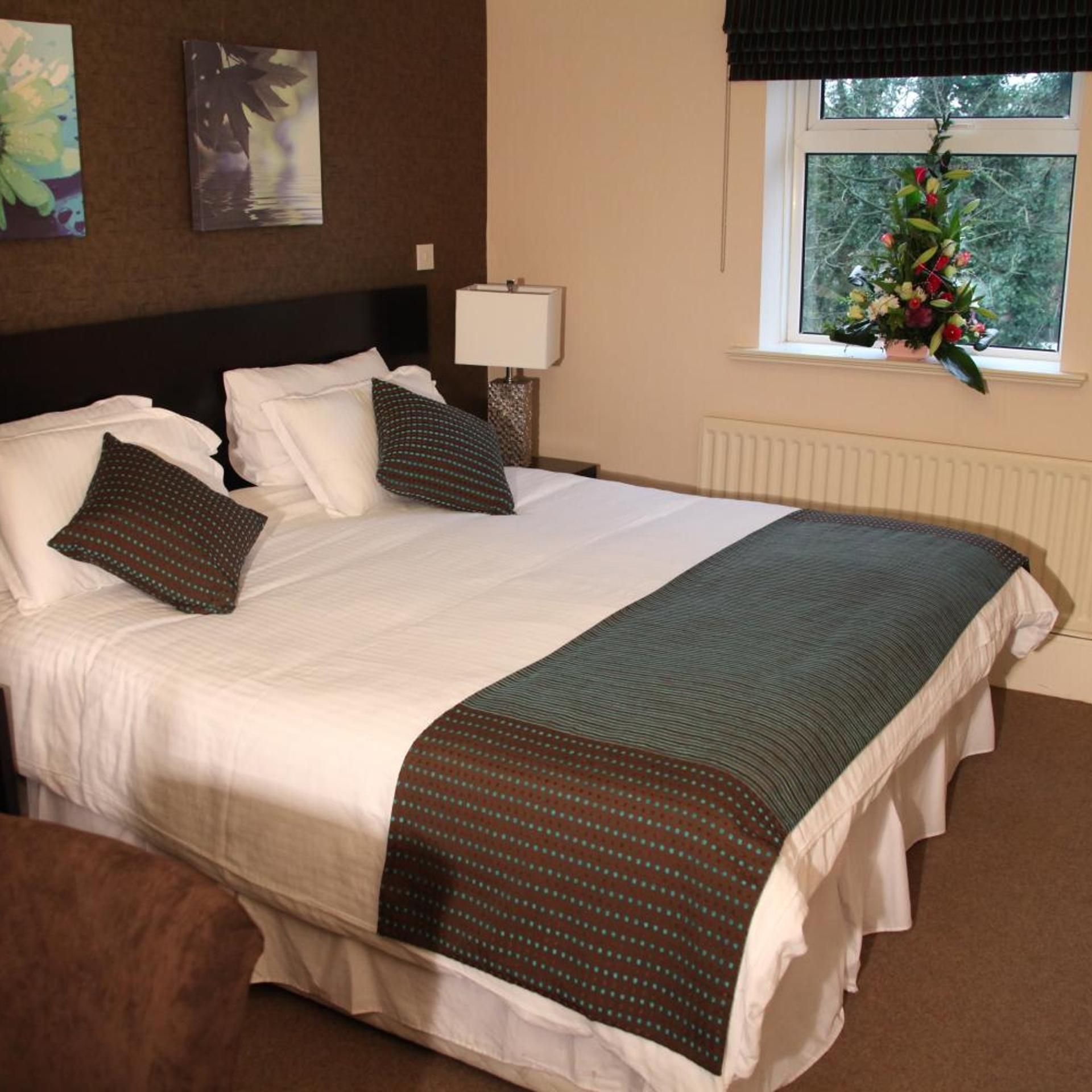 A neatly made bed with white linens and a dark green accent throw, a bedside lamp, a window with a view of greenery, and a wall-mounted artwork.