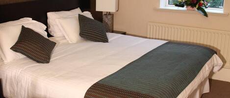 A neatly made bed with white linens and a dark green accent throw, a bedside lamp, a window with a view of greenery, and a wall-mounted artwork.