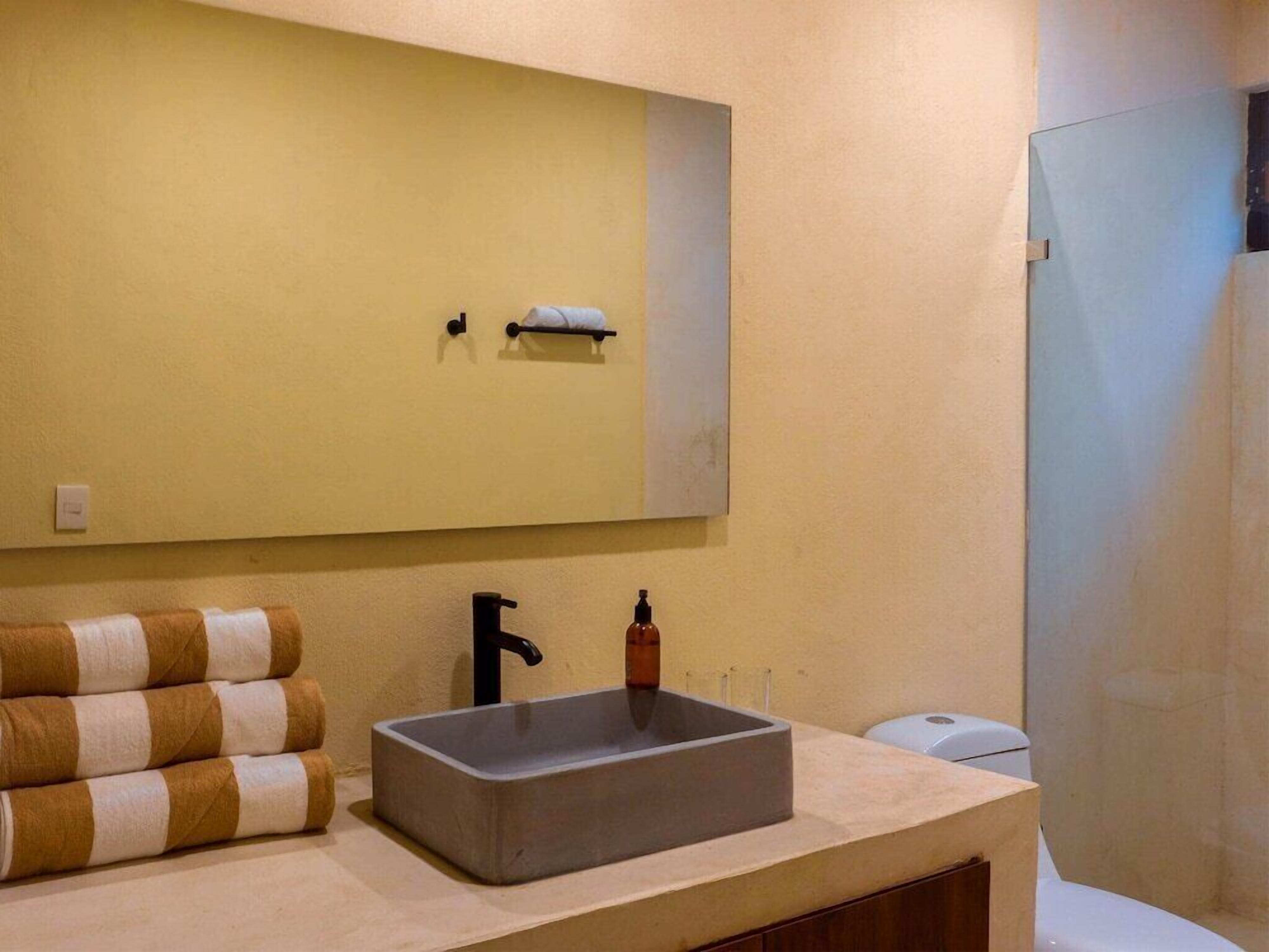Ein Badezimmer mit einem großen Spiegel, einem rechteckigen Waschbecken und einer Toilette. Handtücher sind auf der linken Seite gestapelt.