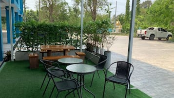 An outdoor patio with tables and chairs, surrounded by greenery and a view of a street.