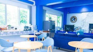 Een moderne kamer met een blauw kleurenschema, met een bank, ronde tafels en een kitchenette met een koelkast en koffieapparaat.