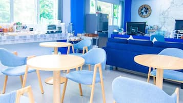 A modern room with a blue color scheme, featuring a sofa, round tables, and a kitchen area with a refrigerator and coffee machine.