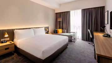 A hotel room with a large bed, bedside table with a lamp, a desk with a chair, and a window with curtains.