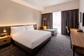 A hotel room with a large bed, bedside table with a lamp, a desk with a chair, and a window with curtains. - Holiday Inn Sepang Airport (Sepang)