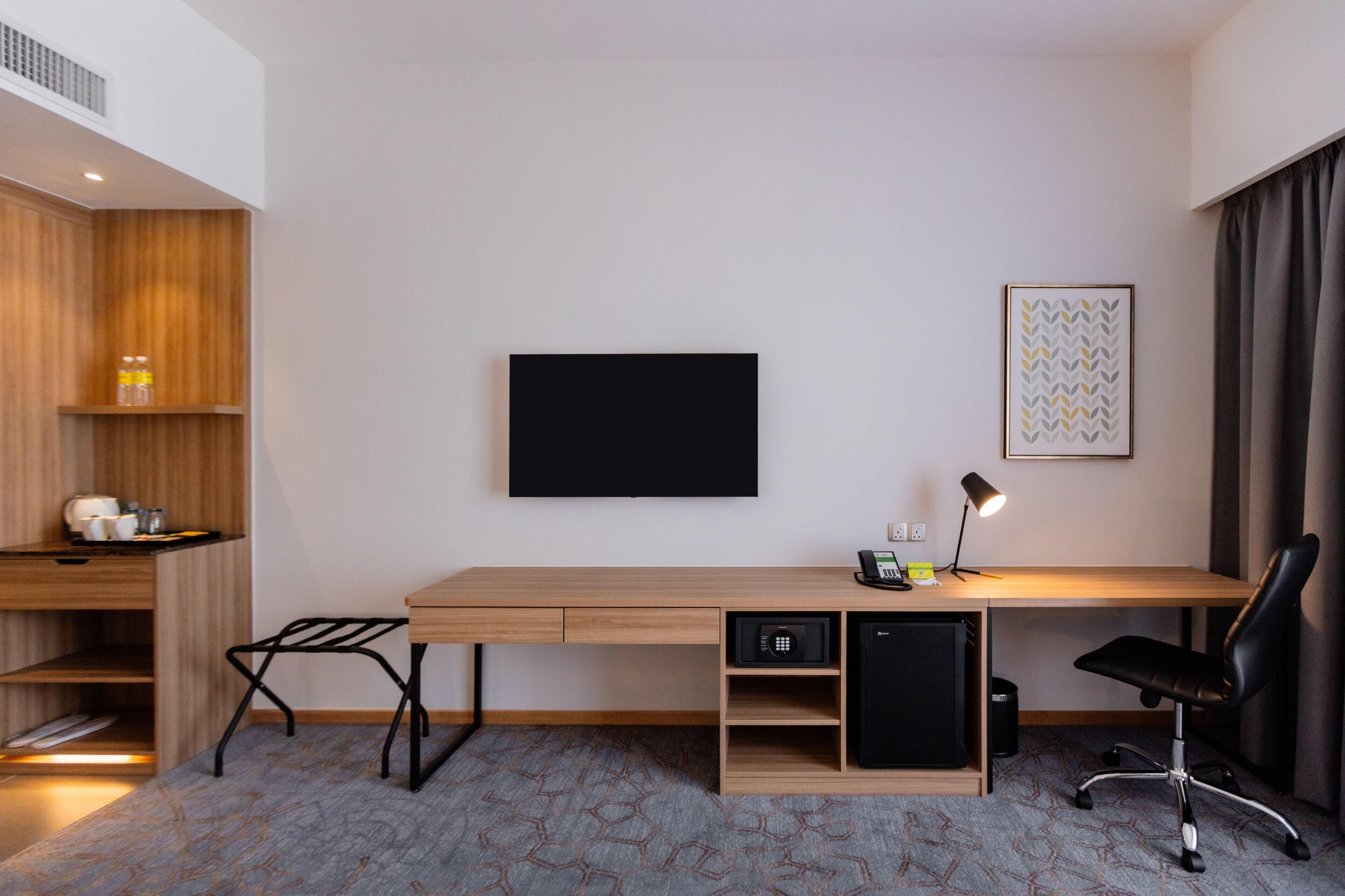 A modern hotel room with a wooden desk, a flat-screen TV, a desk lamp, a black office chair, and a patterned carpet.
