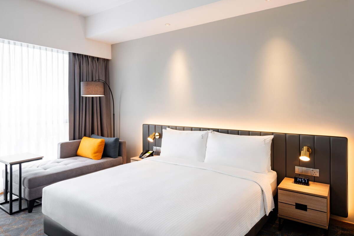 A hotel room with a large bed, a bedside table, a lamp, and a nightstand with a clock.