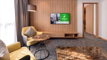 A hotel room with a wooden wall panel, a television mounted above a seating area, and a patterned carpet.