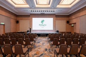 Ein Konferenzraum mit Reihen von Stühlen, einer Bühne mit einem Bildschirm, der das Holiday Inn Logo zeigt, und einem Teppichboden.