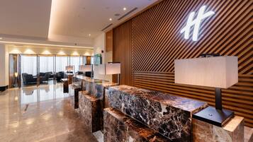 A hotel reception area with a marble counter, wooden panel wall, and a large illuminated 'H' sign.