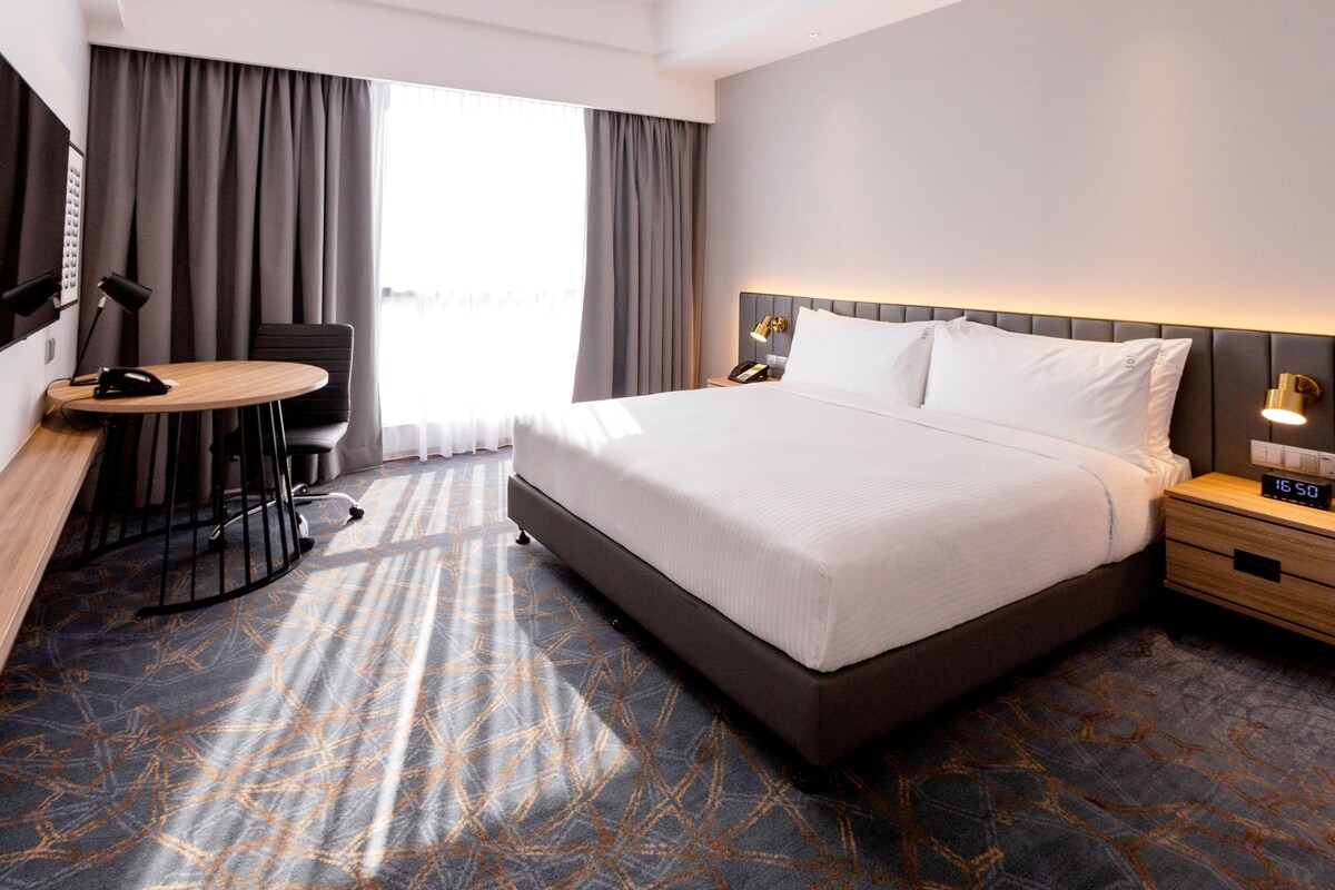 A hotel room with a large bed, a desk with a chair, a nightstand with a lamp, and a view of the outdoors.