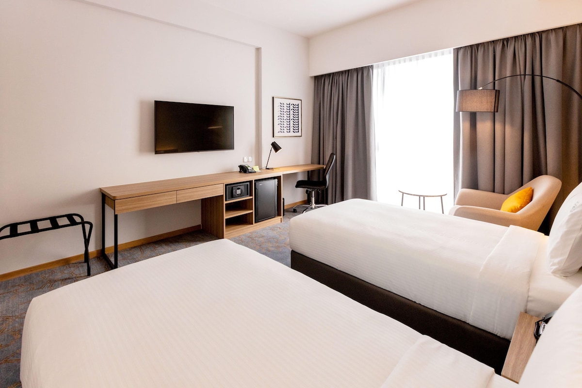 A hotel room with two beds, a desk with a flat-screen TV, a chair, a lamp, and a window with curtains.