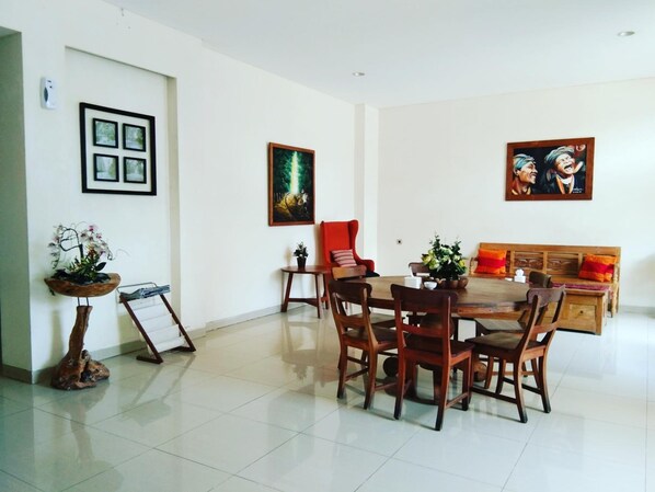 Un comedor con una mesa y sillas de madera, un sillón rojo y un sofá con cojines naranjas. Hay cuadros enmarcados en las paredes y una planta pequeña sobre un soporte.