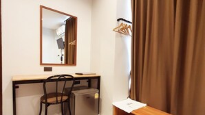 A compact room with a desk, chair, and a small refrigerator. A mirror hangs on the wall, and a wooden shelf is below the window. - Suhotel Suratthani (Surat Thani)