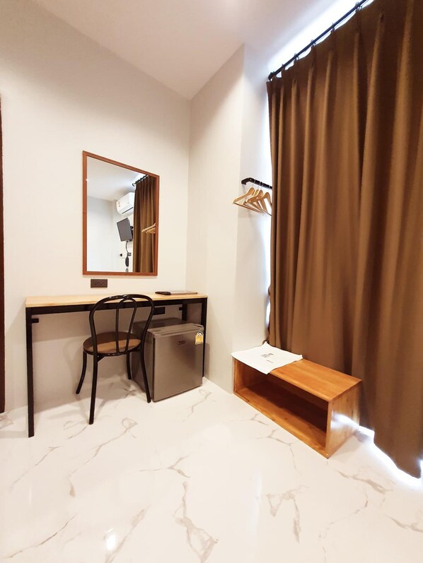 A compact room with a desk, chair, and a small refrigerator. A mirror hangs on the wall, and a wooden shelf is below the window. - Suhotel Suratthani (Surat Thani)