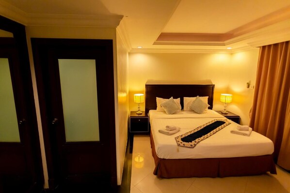 A hotel room with a large bed, two bedside tables with lamps, a headboard, and a door with frosted glass panels. - Ivory Palace Hotel (Pattaya)