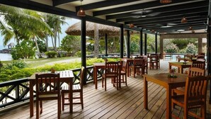 An outdoor dining area with wooden tables and chairs, a thatched roof, and a view of a pool and palm trees. - Waya Island Resort (Yasawa Island)