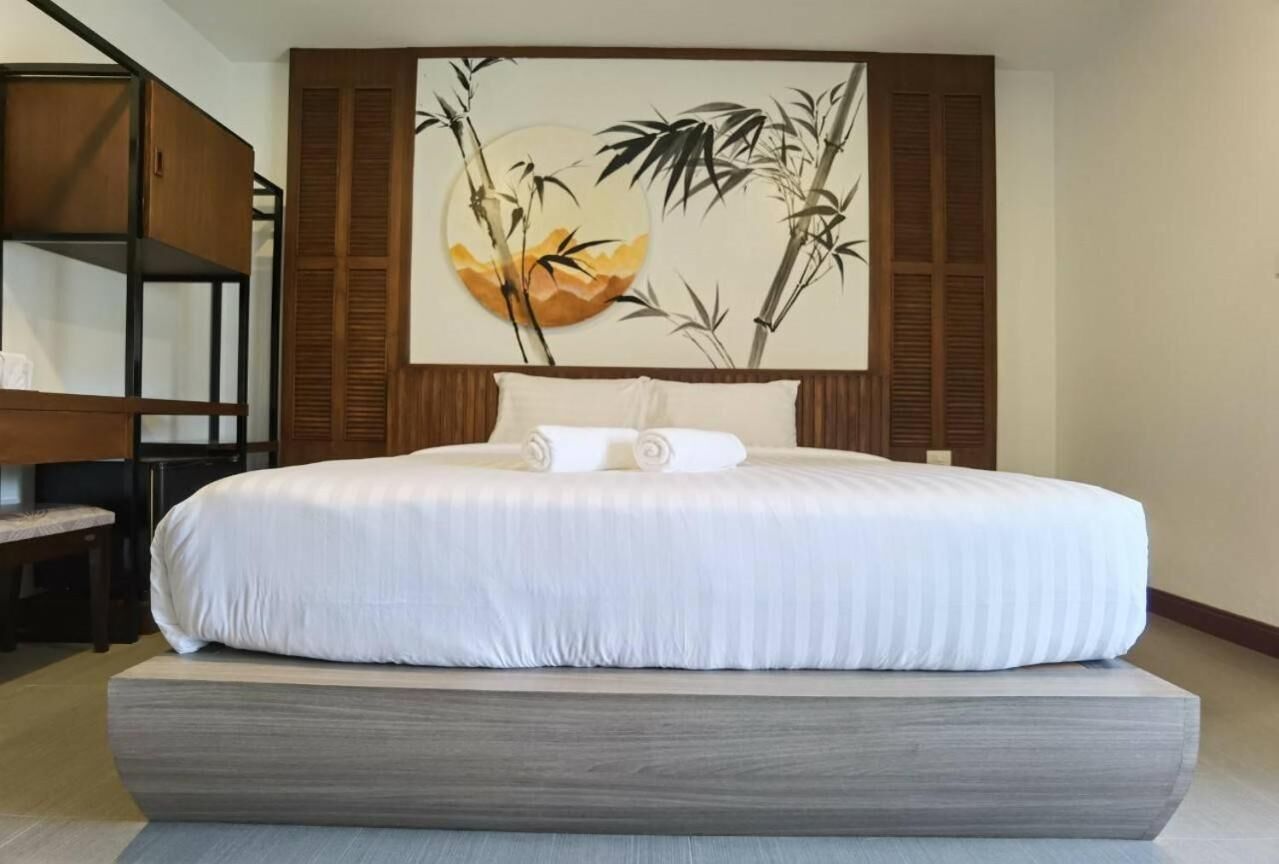 Ein ordentlich bezogenes Bett mit weißer Daunendecke und zwei aufgerollten Handtüchern, einem Holzkopfstück und einem großen Wandbild von Bambus und Blumen.