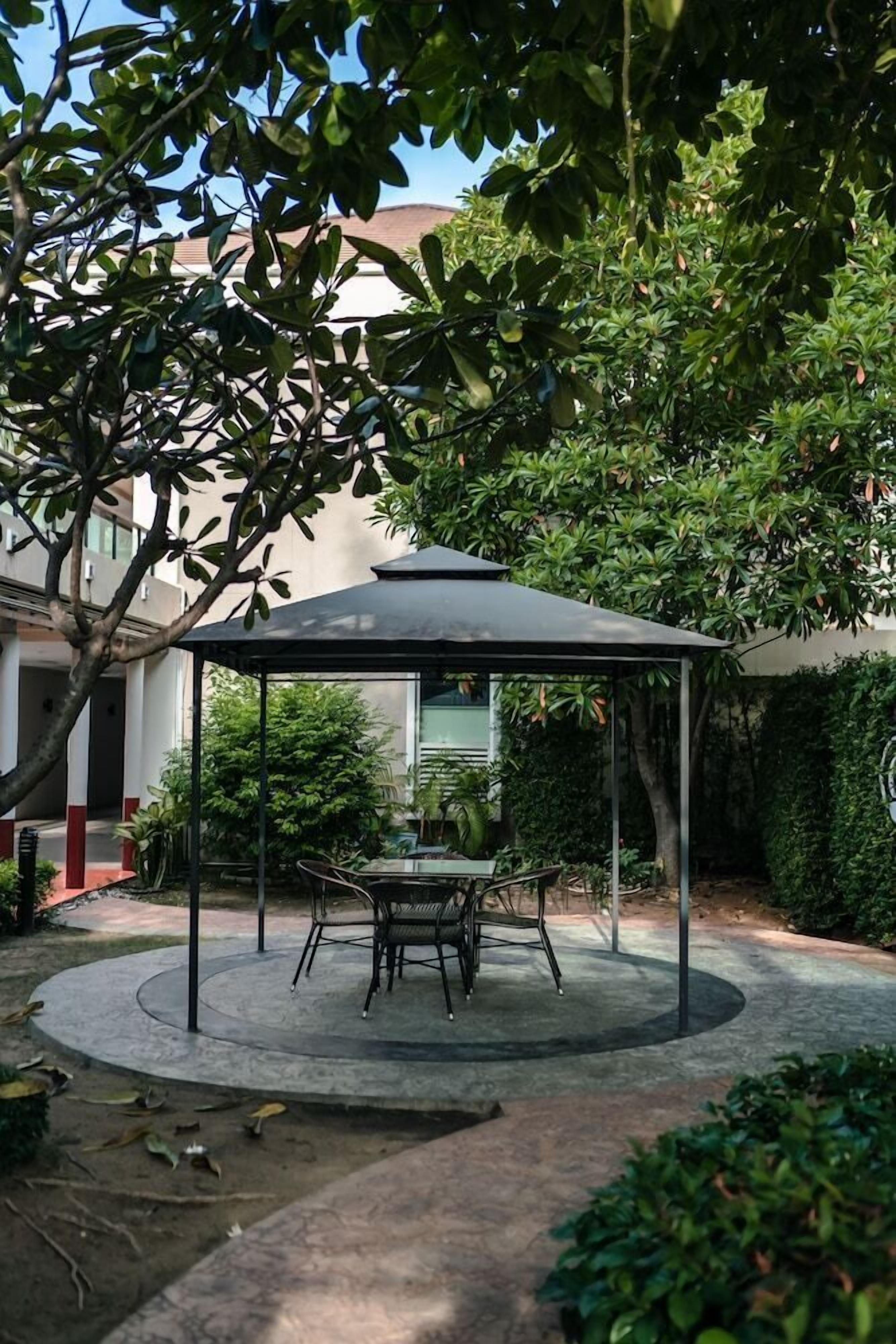 A covered outdoor seating area with a table and chairs, surrounded by greenery and a building in the background.
