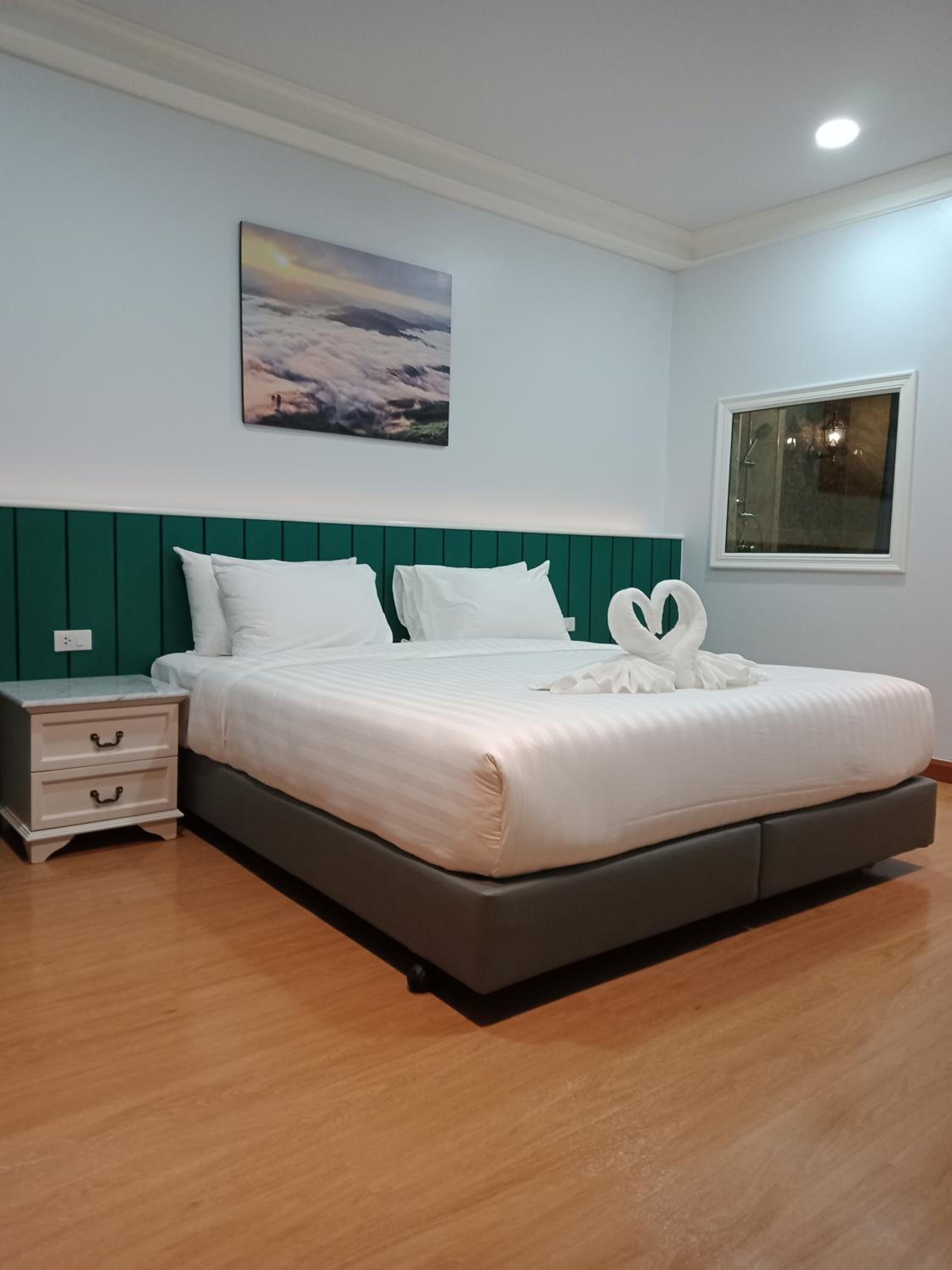 A neatly made bed with white linens and two swan-shaped pillows, a bedside table with drawers, a wall-mounted painting, and a window with a view.