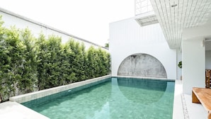 Zona de piscina moderna amb una piscina blava cristal·lina, una banca de fusta i una paret blanca amb un obertura circular.