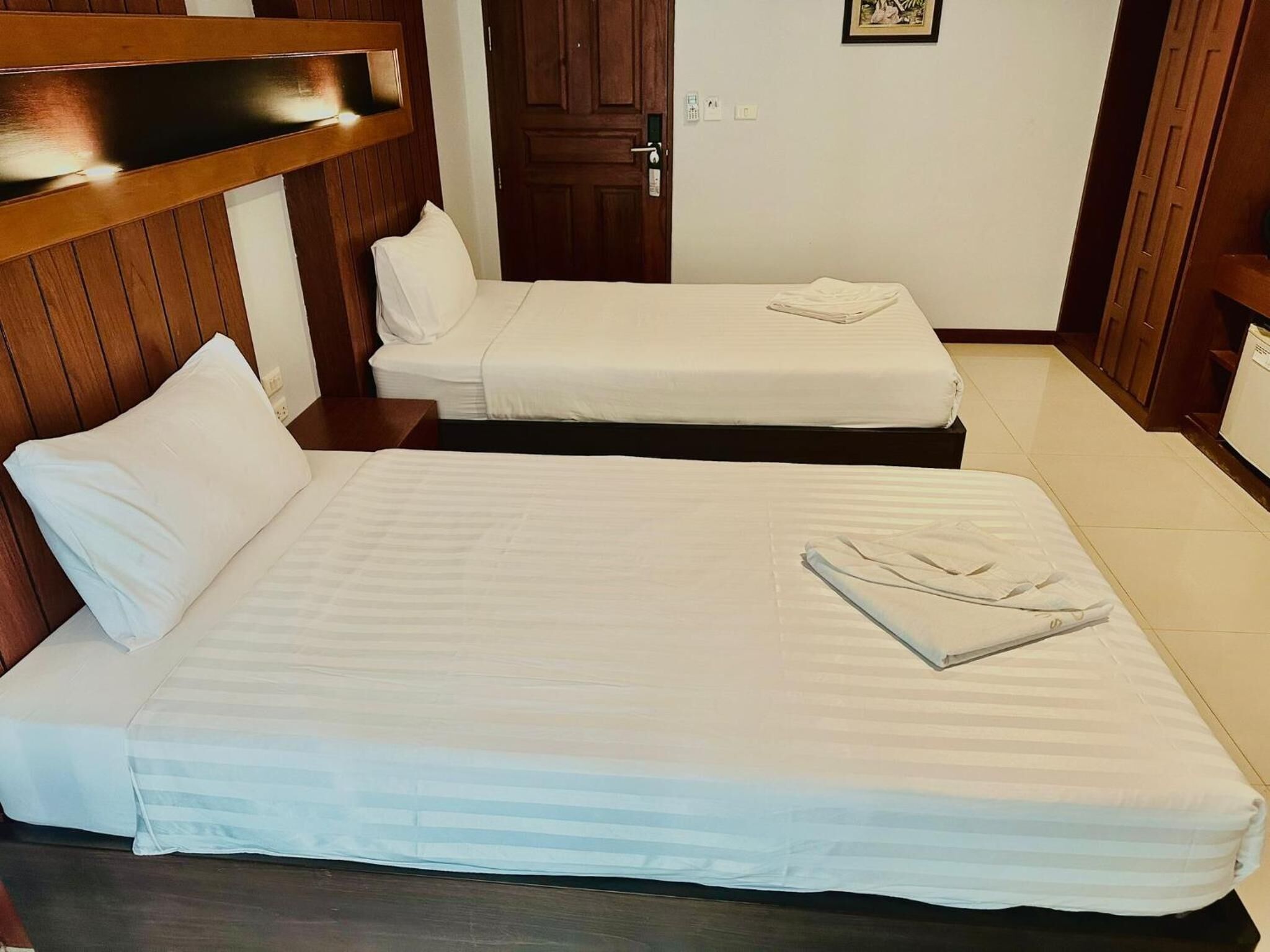 Hotellihuone, jossa on kaksi erillistä sänkyä, puiset päätyosat, ovi ja pieni kehystetty kuva seinällä.