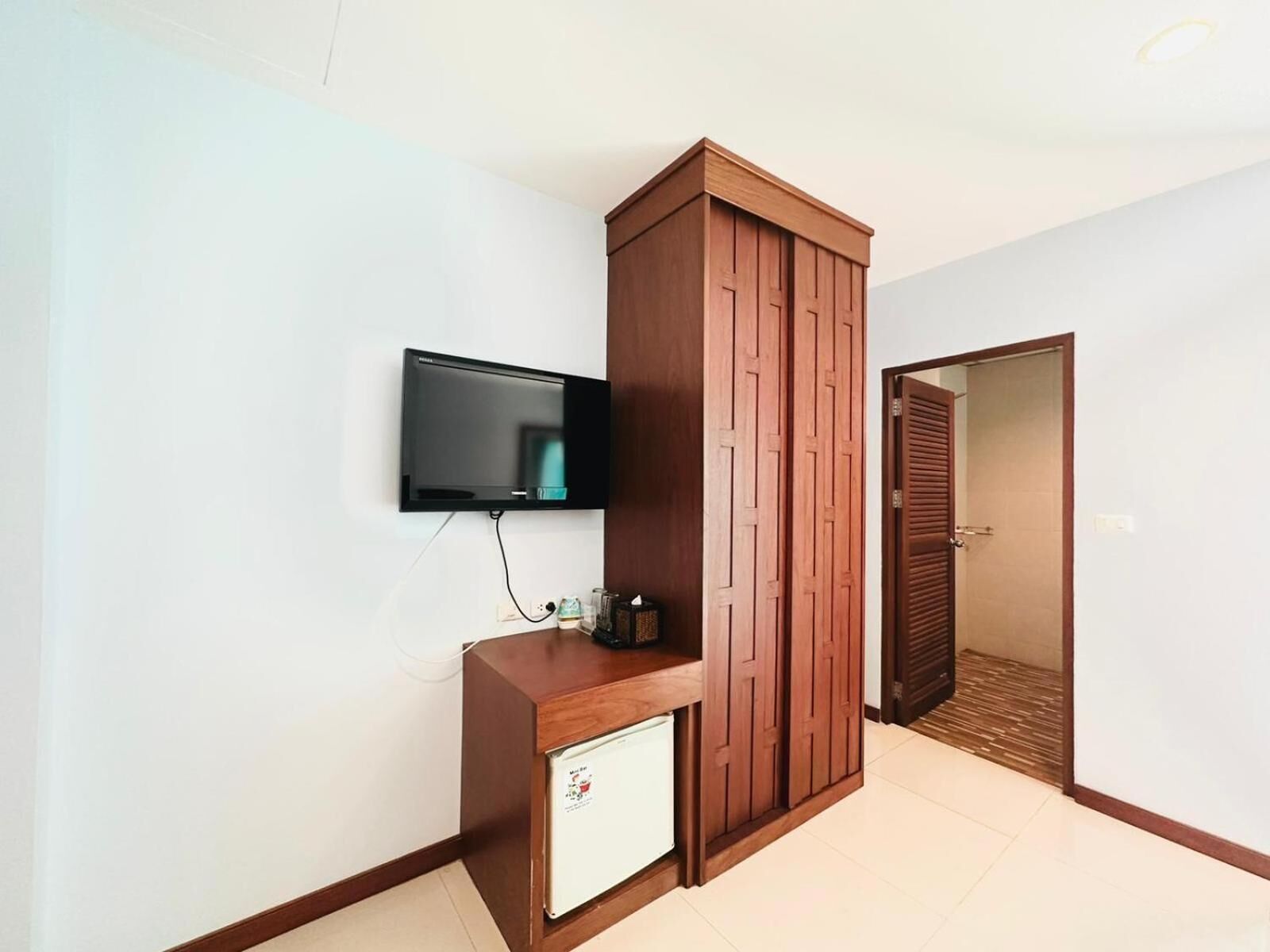 A room with a wooden wardrobe, a small white cabinet, and a mounted television.