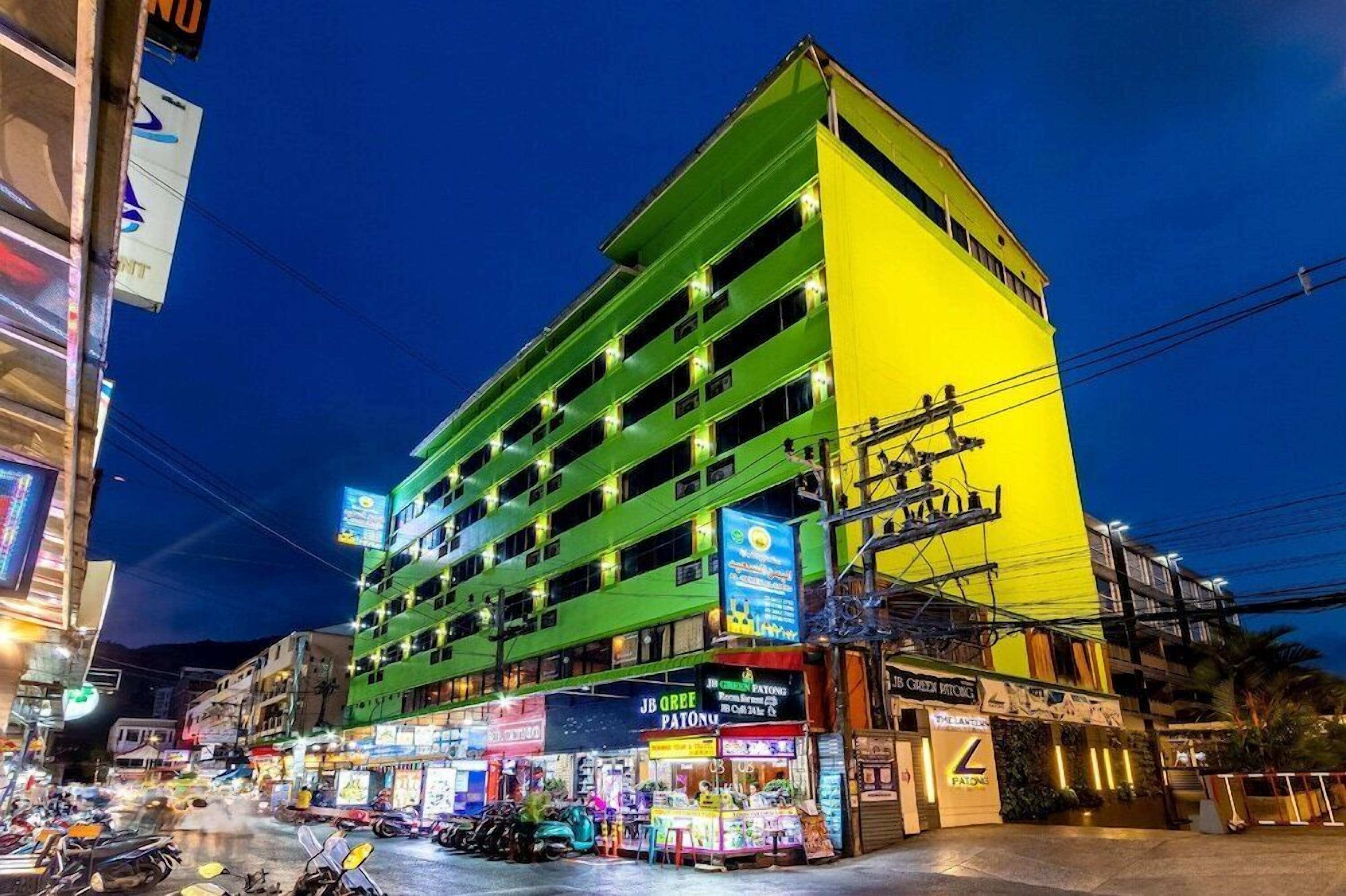 A multi-story building with a green and yellow facade, multiple balconies, and various commercial establishments on the ground floor.